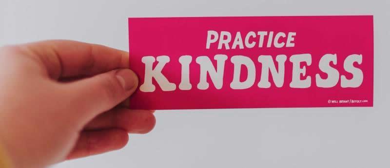Een hand die een roze kaartje vasthoudt met de tekst Practice Kindness, ter illustratie bij het artikel Een andere blik op verpleging